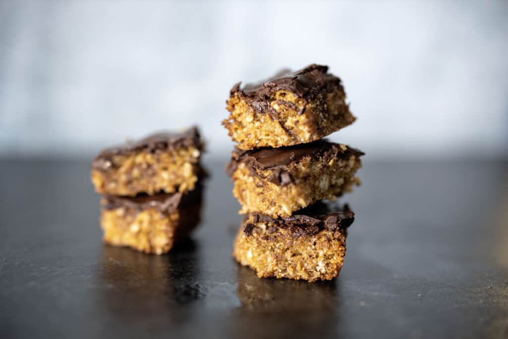 stacked oat bar cookies - golden oat bars with a thin chocolate layer on top. Two cookies stacked to the left with 3 cookies to right side. Lateral view, on black granite