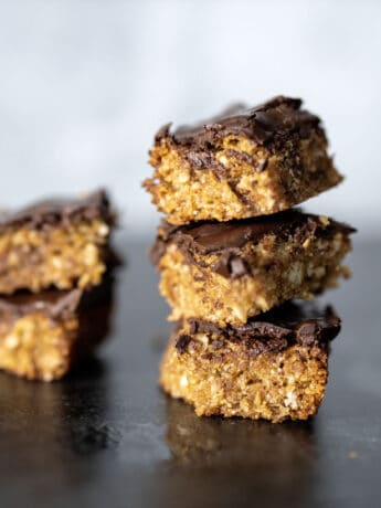 stacked oat bar cookies - golden oat bars with a thin chocolate layer on top. Two cookies stacked to the left with 3 cookies to right side. Lateral view, on black granite