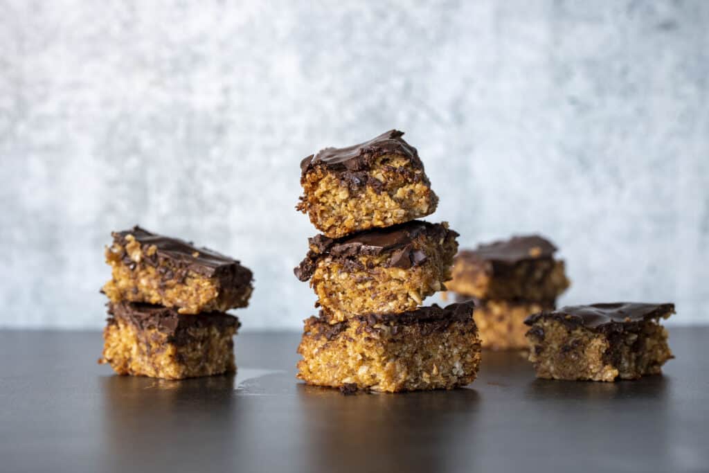 stacked oat bar cookies - golden oat bars with a thin chocolate layer on top. Two cookies stacked to the left with 3 cookies to right side and several more scattered in background.. Lateral view, on black granite