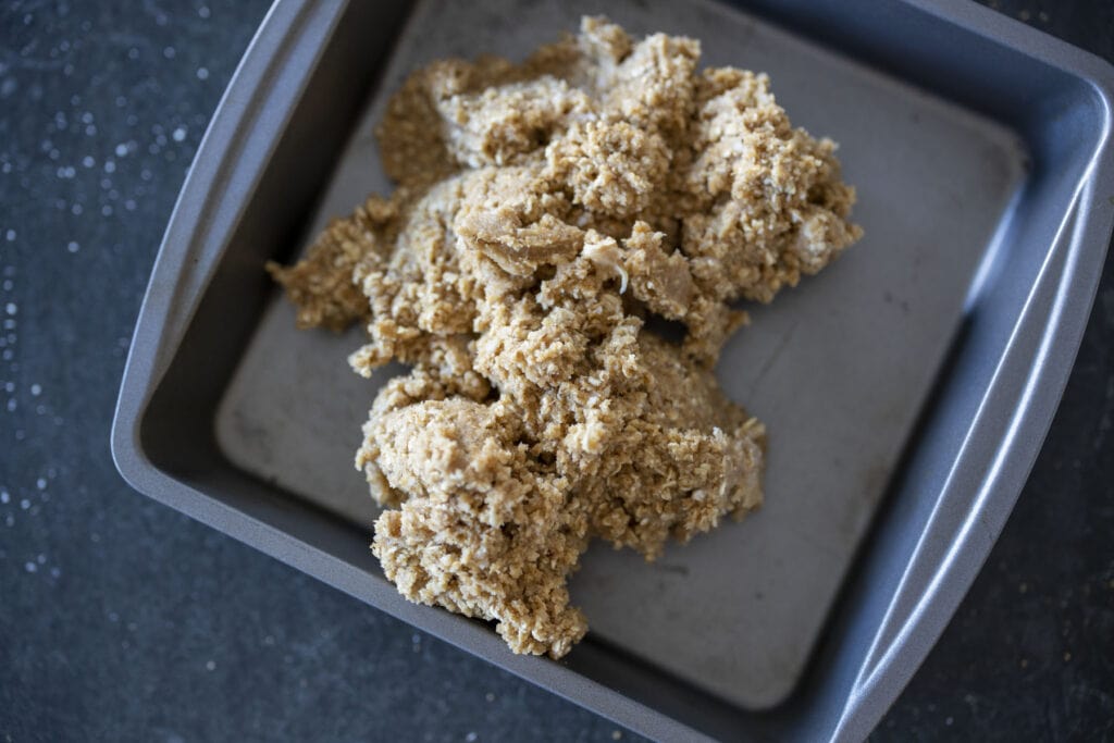 english toffee cookie batter - light brown with visible oats -clumped in center of square tin baking sheet on black granite