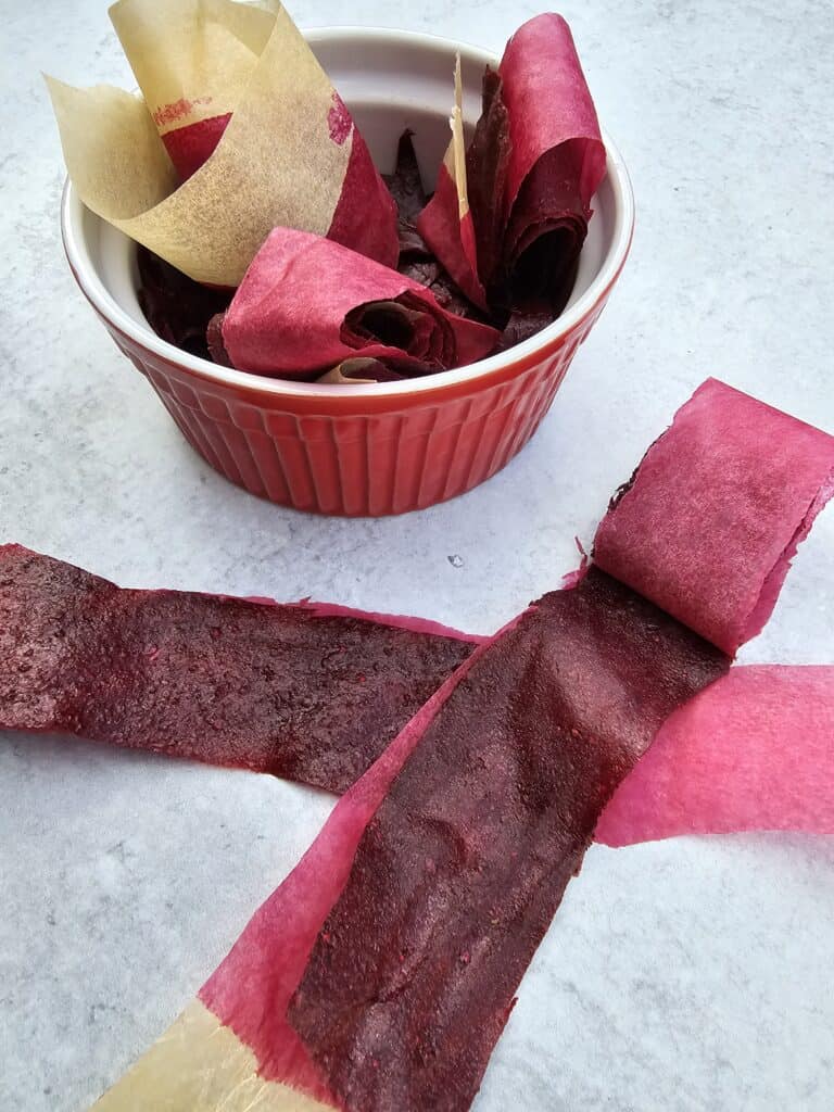 Individual homemade mixed berry fruit roll ups in parchment in a red bowl with two fruit roll ups stretched out on the table in front